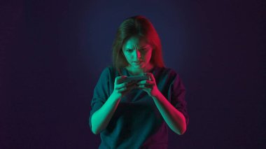 Gergin bir kadın akıllı telefonuyla online oyun oynuyor. Stüdyoda mavi arka planda pembe ve yeşil neon ışıklı bir kadın var.