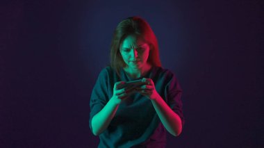 Gergin bir kadın akıllı telefonuyla online oyun oynuyor. Stüdyoda mavi arka planda pembe ve yeşil neon ışıklı bir kadın var.