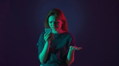 Bir kadın hayal kırıklığıyla akıllı telefon ekranına bakar, kollarını açar. Stüdyoda elinde telefon olan bir kadın. Pembe ve yeşil ışıklı mavi bir arka planda.