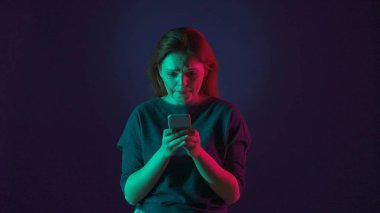 Heyecanlı bir kadın dudaklarını ısırır gergin ve umutsuzca akıllı telefon ekranına bakar. Stüdyoda elinde telefon olan bir kadın. Pembe ve yeşil ışıklı mavi bir arka planda.