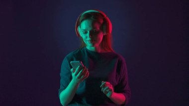 Kırmızı kablosuz kulaklık takan bir kadın müzik dinlemek için akıllı telefon kullanır. Stüdyoda mavi arka planda pembe ve yeşil neon ışıklı bir kadın var.