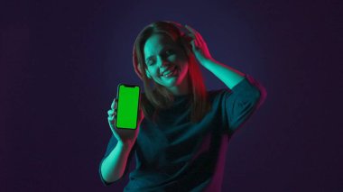 Kırmızı kablosuz kulaklıklı bir kadın yeşil bir akıllı telefon ekranı gösteriyor. Bir kadın, yeşil ve pembe neon ışıklı mavi arka plandaki bir stüdyoda, uygulamayı kullanarak, gözleri kapalı müzik dinler.