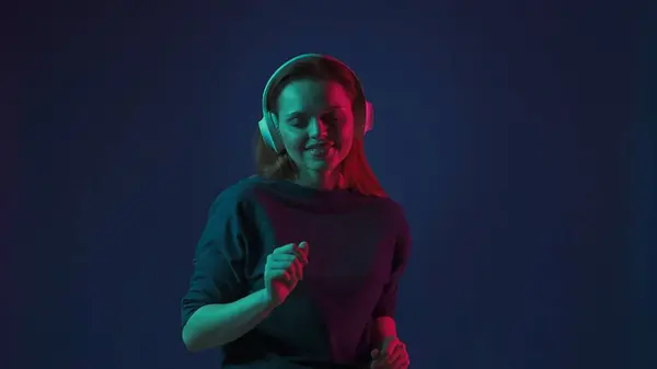 一位头戴白色无线耳机的红头发年轻女子听Dj的音乐和跳舞 粉红和绿色霓虹灯下蓝色背景的工作室里的女人 — 图库照片
