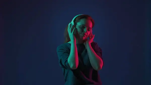 A young redhaired woman wearing white wireless headphones listens to DJ music and dances. Woman in studio on blue background in pink and green neon light