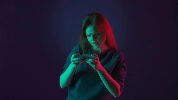 一个紧张的女人在她的智能手机上玩网络游戏 粉红和绿色霓虹灯下蓝色背景的工作室里的女人 — 图库照片