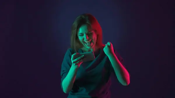 女性はスマートフォンでオンラインゲームを勝ち取った後 勝利ジェスチャーを作ります ピンクと緑のネオンライトの青い背景にスタジオで女性 — ストック写真
