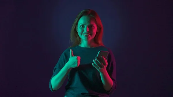 年轻的红头发女人手里拿着智能手机 竖起大拇指 在蓝色背景的演播室里用粉色和绿色霓虹灯照明的女人 — 图库照片