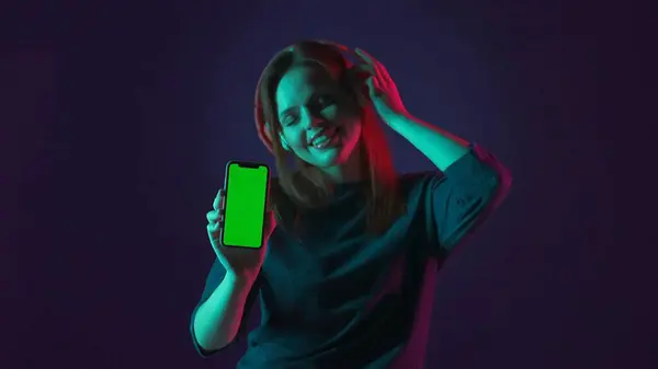 一个戴着红色无线耳机的女人展示了一个绿色的智能手机屏幕 一个女人在一个蓝色背景的工作室里 用一个应用程序 用绿色和粉色霓虹灯 闭着眼睛听音乐 — 图库照片