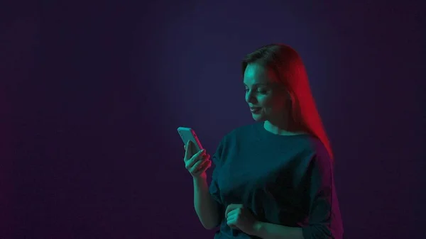 笑顔の若い赤毛の女性は スマートフォンを手に持って ビデオ ソーシャルネットワークを見て メールをチェックします スタジオでピンクと緑のネオンライトで照らされた女性 — ストック写真