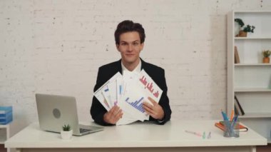 Ofiste oturan genç bir adamın elinde grafikleri ve çizelgeleri gösteren orta büyüklükte bir videosu. Adam mutlu ve memnun bir şekilde kağıtları havaya fırlatıyor. İş, kariyer büyümesi, reklam.