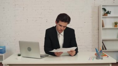 Ofiste oturan genç bir adamın masasındaki grafikleri ve çizelgeleri kontrol ederken çekilmiş orta boy videosu. Kafası karışmış görünüyor, ellerini açıyor, omuz silkiyor. İş, kariyer, reklam.