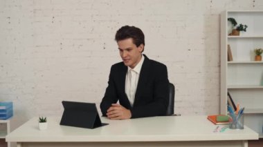 Ofiste, tabletinin önünde oturmuş video aramasını bekleyen genç bir adamın orta boy videosu. Sonra konuşmaya başlıyor, biriyle konuşuyor, mutlu ve pozitif. İş, web kaynakları, uygulama.