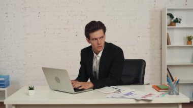 Ofiste oturan bir gencin orta boy videosu, dizüstü bilgisayarla çalışıyor, yan tarafa bakıyor ve bir şeyler düşünüyor. Not alıyor, bir şeyler yazıyor. İş ilanı, resmi tarz..