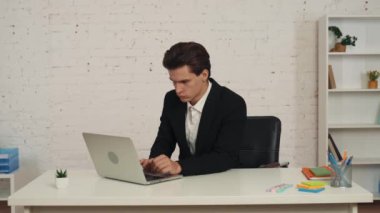 Masada oturan genç bir adamın dizüstü bilgisayarla çalışırken çekilmiş orta boy videosu. Kameraya bakıyor, rahat ve kendinden emin. İş, resmi tarz, istihdam, reklam.