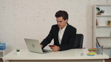 Masada dizüstü bilgisayar kullanan ve elinde kredi kartı olan genç bir adamın internetten alışveriş yaparken çekilmiş orta boy videosu. O, kameraya güvenle bakar. Web sitesi ilanı, online satın alma.