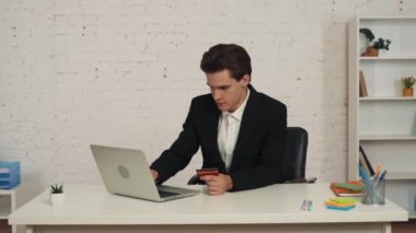 Masada dizüstü bilgisayar kullanan ve elinde kredi kartı olan genç bir adamın internetten alışveriş yaparken çekilmiş orta boy videosu. Teslimat paketleri alıyor. Teslimat hizmeti, web sitesi, çevrimiçi alım.