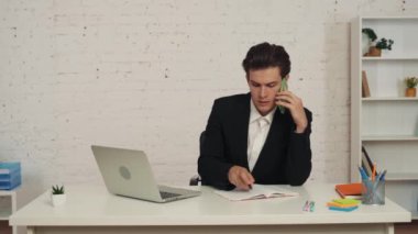 Masada oturan genç bir adamın orta boy videosu, iş görüşmesi, telefon görüşmesi, birileriyle konuşma, bir şeyler yazma, not alma. İş, anlaşma, ofis, istihdam.