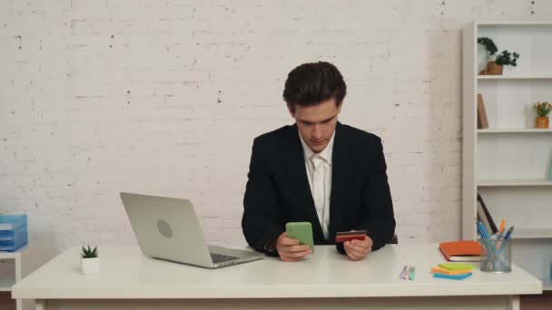 一个年轻人坐在桌旁 拿着智能手机和信用卡 完成网上购物的中等视频 他自信地看着摄像机 App Website Advertising Online Purchase — 图库视频影像