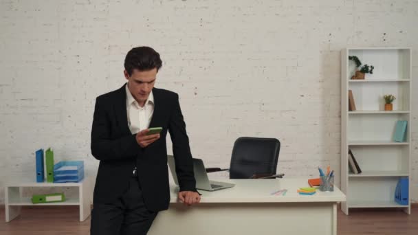 一个年轻人在办公室里 手里拿着智能手机 收到消息 被告知坏消息的中等视频 他的反应是沮丧和失望 失败的交易 — 图库视频影像
