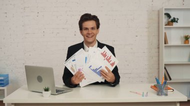 Ofiste oturan genç bir adamın elinde grafikleri ve çizelgeleri gösteren orta boy bir fotoğrafı. Adam kameraya bakıyor, mutlu ve memnun. İş, kariyer büyümesi, reklam.