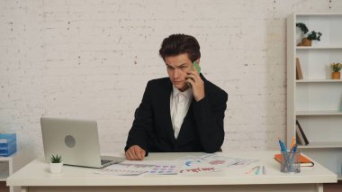 Masada oturan genç bir adamın orta boy fotoğrafı, iş görüşmesi, telefon görüşmesi, birileriyle konuşma ve yan tarafa bakma, odaklanmış ve konsantre. İş, anlaşma, ofis, istihdam