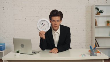 Ofisteki masada oturan, elinde saatle kameraya bakan, uykulu, yorgun ve bitkin bir gencin orta boy görüntüsü. İş, aşırı çalışma, yerleşik yaşam tarzı.