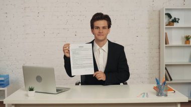 Ofisteki masada oturan genç bir adamın elinde bir kontrat tutarken, yüzünde gururlu bir ifadeyle, teşvik edici ve davetkar bir şekilde görüntüsü. İş teklifi, reklam..