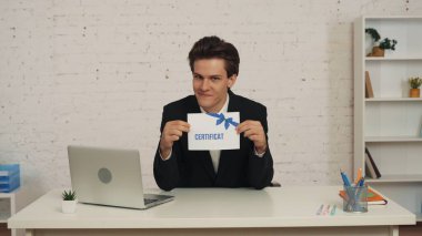 Ofisteki masada oturan genç bir adamın, elinde sertifikayla, yüzünde gururlu bir ifadeyle, teşvik edici ve davetkar bir şekilde oturduğu orta ölçekli bir fotoğraf. İş, teklif, kurslar.