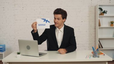 Ofisteki masada oturan genç bir adamın, elinde sertifikayla, yüzünde gururlu bir ifadeyle, teşvik edici ve davetkar bir şekilde oturduğu orta ölçekli bir fotoğraf. İş, teklif, kurslar.