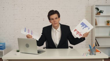 Ofisteki masada oturan genç bir adamın grafikleri ve çizelgeleri gösterirken orta boy fotoğrafı. Kameraya bakıyor, onaylamıyor ve tatmin olmuyor. İş yerinde sorun, beyin fırtınası, problem çözme.