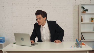 Ofiste çağrı merkezi kulaklığı takan, biriyle konuşan, bilgisayarında çalışan, yardım eden, destek veren bir gencin orta boy fotoğrafı. İş ve çağrı merkezleri, ajanslar için reklam.