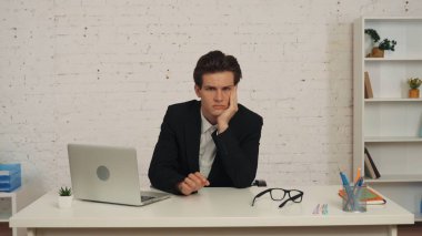 Ofisteki masada oturan genç bir adamın orta boy fotoğrafı, işe ara verirken, eline yaslanırken, yorgun, yorgun ya da sıkılmış. İş ilanı, istihdam.