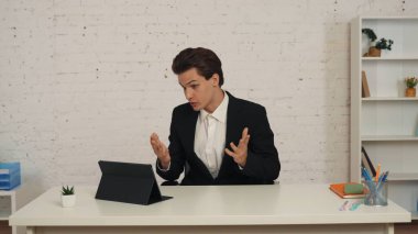 Ofiste, tabletinin önünde oturmuş video aramasını bekleyen genç bir adamın orta boy fotoğrafı. Sonra konuşmaya başlıyor, biriyle konuşuyor, mutlu ve pozitif. İş, web kaynakları, uygulama.