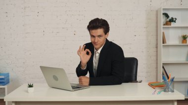 Ofisteki masada oturan, dizüstü bilgisayarının başında çalışan bir gencin orta boy fotoğrafı. Tamam işareti, jest, bir şey öneriyor. İş fırsatı, iş ilanı.