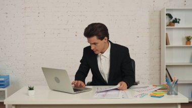 Ofiste oturan genç bir adamın orta boy fotoğrafı, bir dizüstü bilgisayar üzerinde çalışıyor ve masasındaki grafikleri ve çizelgeleri kontrol ediyor. Odaklanmış ve konsantre olmuş görünüyor. İş ilanı, istihdam, araştırma.