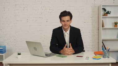 Masada oturan genç bir adamın orta boy fotoğrafı, parmakları birbirine kenetlenmiş, birbirine kenetlenmiş, özgüven ve kararlılığını ifade eden. İş ilanı, istihdam, bayilik.