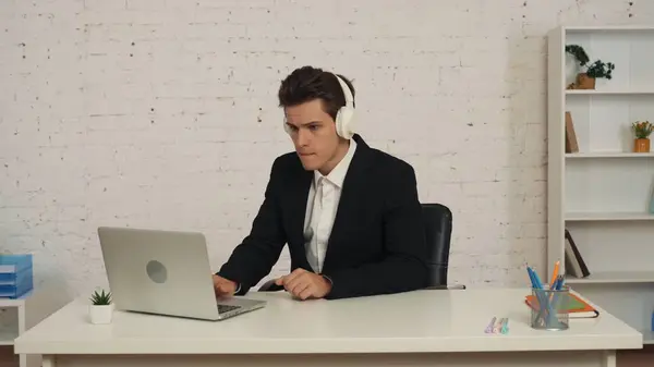 一个年轻人在办公室里工作 在笔记本电脑前 通过耳机听音乐 坐在椅子上 拍下了一张中等的照片 音乐平台 设备和技术 — 图库照片
