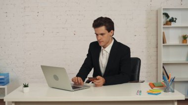 Masada oturan genç bir adamın elinde akıllı telefon ve kredi kartıyla internet üzerinden ödeme yaparken orta boy bir fotoğrafı. Uygulama, web sitesi reklamı, online alışveriş, satın alma.