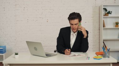 Genç bir adamın masada otururken, iş görüşmesi yaparken, telefonla konuşurken, birileriyle konuşurken, bir şeyler yazarken, not alırken orta boy bir fotoğraf. İş, anlaşma, ofis, istihdam.