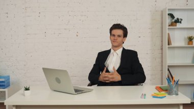 Masada oturan genç bir adamın orta boy fotoğrafı, parmakları birbirine kenetlenmiş, birbirine kenetlenmiş, özgüven ve kararlılığını ifade eden. İş ilanı, istihdam, bayilik.