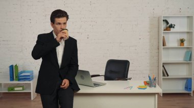 Genç bir adamın çalışma alanının, masasının, içkisinin, içkisinin, kahvesini yudumlarken ve düşünceli bir şekilde yan tarafına bakarken orta boy fotoğrafı. İşsizlik, resmi tarz, istihdam, reklam.