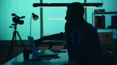 Bir dedektifin orta boy siluet videosu, sorgu odasında oturan, sigara içen, dizüstü bilgisayarda çalışan ve telefonda konuşan bir polis. Gerçek suç, belgesel, yaratıcı içerik.