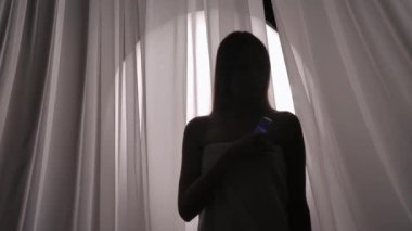 Havluya sarılı bir kadın siluetinin orta görüş videosu. Yüzüne sprey sıkılmış. Boğuk bir ışıkta cildine nemlendirici bir cihaz takılmış. Ürün reklamı, kişisel bakım rutini.