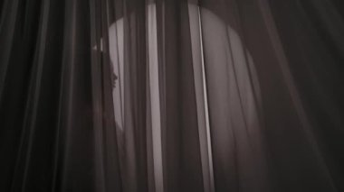 Perdenin arkasındaki çerçeveye sarılı bir kadın siluetinin orta boy videosu. Boğuk bir ışıkta saçlarını okşuyor, okşuyor. Ürün reklamı, kişisel bakım rutini.