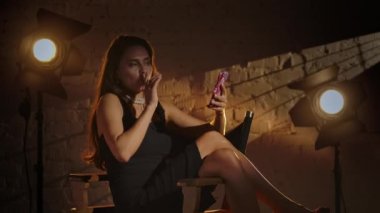 Sinematografi ve sahne arkası yaratıcı konsepti. Siyah elbiseli çekici esmer kadın ve inciler yönetmen koltuğunda oturuyor, lolipop tutuyor ve akıllı telefondan selfie çekiyor..