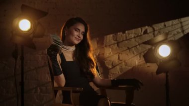 Sinematografi ve sahne arkası yaratıcı konsepti. Siyah elbiseli zarif esmer kadın, ipek eldivenler ve inciler yönetmen koltuğunda oturuyor, elinde para fanı ile gülümsüyor..