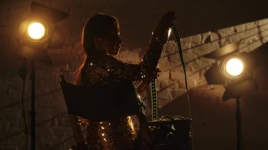 Sinematografi ve sahne arkası yaratıcı konsepti. Altın elbiseli zarif esmer kadının silueti yönetmen koltuğunda oturuyor, uzun bir film şeridi tutuyor ve ona bakıyor..