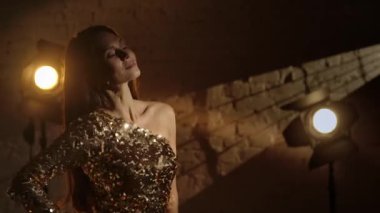 Sinematografi ve sahne arkası yaratıcı konsepti. Çekici, altın elbiseli esmer bir kadının kareye doğru yürüdüğü ve fotoşopa poz verdiği yakın plan, etrafta el fenerleri, gülümseyen.