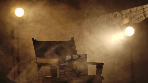 シネマトグラフィーと映画の舞台は 創造的なコンセプトを舞台にしています 監督の椅子に立っている黒いクラッパーボードのクローズアップショット バックグラウンドのプロジェクター 暖かい光と煙が動き回る — ストック動画