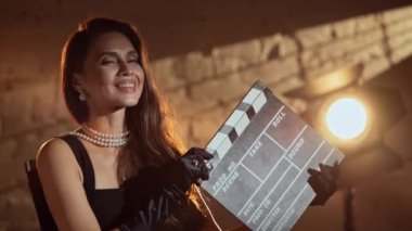 Sinematografi ve sahne arkası yaratıcı konsepti. Siyah elbiseli, ipek eldivenli, yönetmen koltuğunda oturan, elinde klaket olan ve kameraya gülümseyen bir kadın resmi..